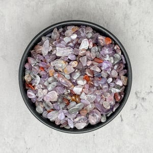 May include: A black bowl filled with a variety of small, polished gemstones. The stones are mainly shades of purple, with accents of red, orange, and clear. The bowl is round and sits on a light grey surface.