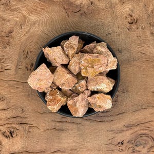 Puede incluir: Un cuenco negro lleno de rocas ásperas de color marrón rojizo. Las rocas tienen una superficie texturizada y diferentes tonos de marrón y beige. El cuenco está sobre una superficie de madera con un patrón de vetas natural.