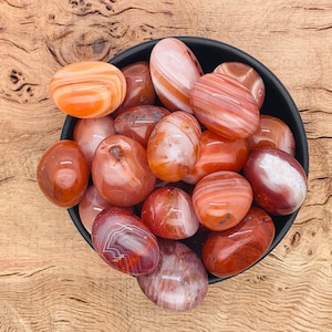 Op de afbeelding: Een kom met getrommelde carneolstenen. De stenen zijn een mix van oranje, rood en wit.