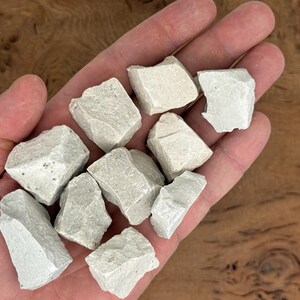 Puede incluir: Primer plano de una mano que sostiene varias rocas de color gris claro, de forma irregular. Las rocas tienen una superficie rugosa y texturizada. El fondo es una superficie de madera borrosa.