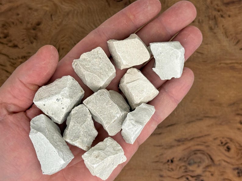 Puede incluir: Primer plano de una mano que sostiene varias rocas de color gris claro, de forma irregular. Las rocas tienen una superficie rugosa y texturizada. El fondo es una superficie de madera borrosa.