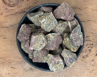 May include: A black bowl filled with rough, raw stones. The stones are primarily a deep reddish-purple color with patches of green and gray. The bowl sits on a wooden surface.
