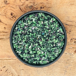 Może przedstawiać: Mała czarna miseczka wypełniona zielonymi, czarnymi i białymi kawałkami kamieni szlachetnych. Miseczka stoi na drewnianej powierzchni.