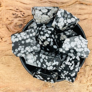 Op de afbeelding: Een kom met zwarte en witte snowflake obsidian getrommelde stenen. De stenen zijn glad en gepolijst, met een gevlekt patroon van wit op zwart.