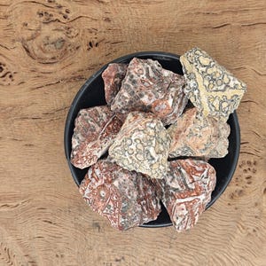 以下が含まれることがあります： 赤い、茶色、白の模様の石が、小さな黒いボウルに入っています。石は独特な大理石のような外観をしています。