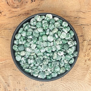 May include: A black bowl filled with green and white speckled stones. The stones are smooth and rounded, varying in size. The bowl rests on a light brown, textured wooden surface. The stones are likely decorative.