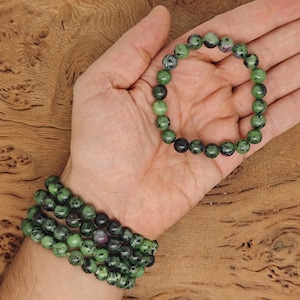 May include: Two green and black gemstone bracelets with round beads. The bracelets are stacked on a person's wrist.
