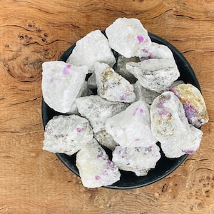 May include: A bowl of white rough ruby crystals with pink and purple inclusions. The crystals are in a black bowl on a wooden surface.