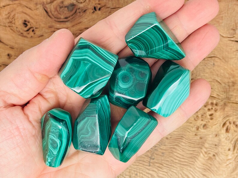 May include: A collection of polished malachite stones, each displaying a rich green color with intricate, swirling patterns. The stones vary in size and shape, showcasing the natural beauty of the mineral. The malachite is held in a hand.