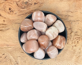 May include: A bowl of tumbled sunstone crystals. The stones are a light peach color with white streaks.
