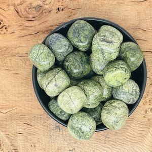May include: A bowl of green serpentine tumbled stones. The stones are smooth and polished, with a variety of green shades and patterns.