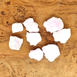 May include: A collection of light pink, rough-cut gemstones arranged on a textured, light brown wooden surface. The gemstones vary in size and shape, with a slightly translucent appearance. The wood grain adds a natural, earthy contrast to the stones.