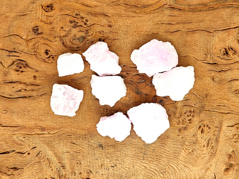 May include: A collection of light pink, rough-cut gemstones arranged on a textured, light brown wooden surface. The gemstones vary in size and shape, with a slightly translucent appearance. The wood grain adds a natural, earthy contrast to the stones.