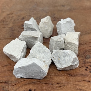 Puede incluir: Un conjunto de rocas de color gris claro, de forma irregular. Las piedras tienen una superficie rugosa y texturizada y var&iacute;an en tama&ntilde;o. Est&aacute;n dispuestas en un mont&oacute;n sobre una superficie de madera, creando una est&eacute;tica natural y terrosa.