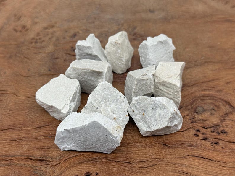 Puede incluir: Un conjunto de rocas de color gris claro, de forma irregular. Las piedras tienen una superficie rugosa y texturizada y var&iacute;an en tama&ntilde;o. Est&aacute;n dispuestas en un mont&oacute;n sobre una superficie de madera, creando una est&eacute;tica natural y terrosa.