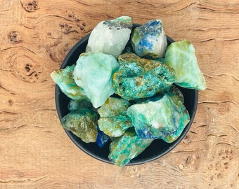 May include: A bowl of green and blue rough gemstones. The stones are various shades of green and blue, with some having a white or clear appearance.