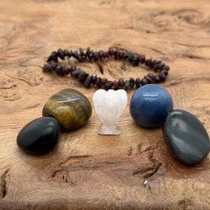 Op de afbeelding: Een verzameling kristallen en edelstenen, waaronder een donkerrode kralenarmband, een tijgeroogsteen, een zwarte steen, een wit engelenbeeldje, een blauwe bol en een grijze steen. De items zijn gerangschikt op een houten oppervlak.