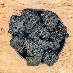 Peut inclure: Un bol noir rempli de morceaux de roche volcanique gris foncé. Les roches ont une texture rugueuse et poreuse.