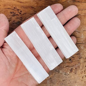 K&ouml;nnte beinhalten: Drei wei&szlig;e Selenit-St&auml;be, auch bekannt als Selenit-St&auml;be, sind in einer Reihe auf einer Hand angeordnet. Die St&auml;be sind glatt und haben eine nat&uuml;rliche, gestreifte Textur.