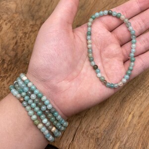 May include: Several beaded bracelets in shades of green, blue, and brown. One bracelet is worn on the wrist, while another is held in the palm of a hand. The small, round beads are strung together to form the bracelets.