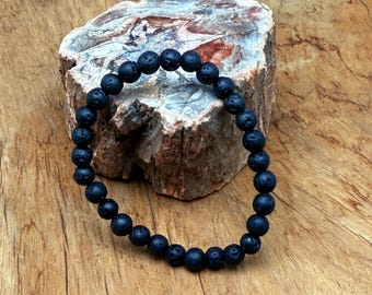 Puede incluir: Una pulsera de piedra de lava negra con cuentas redondas. La pulsera está ensartada y descansa sobre un trozo de piedra rugosa de color claro. El fondo es una superficie de madera.