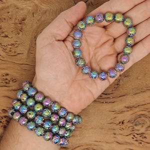 May include: Two bracelets made of iridescent lava rock beads. The bracelets are stacked on a person's wrist. The beads are round and have a textured surface. The bracelets are a dark gray color with hints of purple, blue, and green.