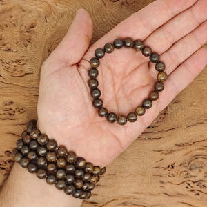 Puede incluir: Un brazalete de madera marrón con cuentas redondas de color marrón oscuro. El brazalete está hecho de cuentas redondas y se muestra en la mano de una persona.