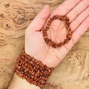 Puede incluir: Dos pulseras hechas de pequeñas piedras irregulares de color marrón rojizo. Las pulseras están apiladas en la muñeca de una persona. La pulsera de arriba es más pequeña y se sostiene en la mano de la persona.