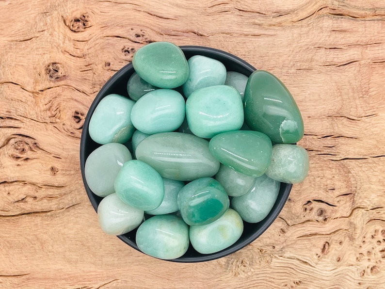 May include: A bowl filled with smooth, light green tumbled stones. The stones are various sizes and shapes, and they are arranged in a random pattern. The bowl is black and has a glossy finish.