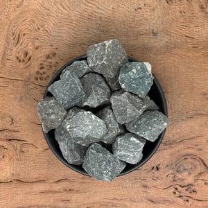 Peut inclure: Un bol noir rempli de pierres rugueuses gris-vert. Les pierres ont une surface texturée et des nuances de gris et de vert. Le bol repose sur une surface en bois avec un motif de grain naturel.