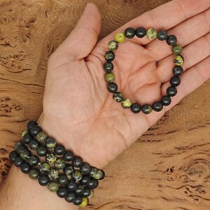 May include: Two beaded bracelets. One bracelet is worn on a wrist, and the other is held in a hand. The bracelets are made of dark gray and olive green beads. The background is a wooden surface.