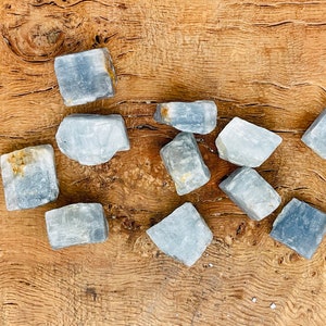 May include: A collection of ten blue calcite crystals, each with a unique shape and texture. The crystals are arranged on a wooden surface.