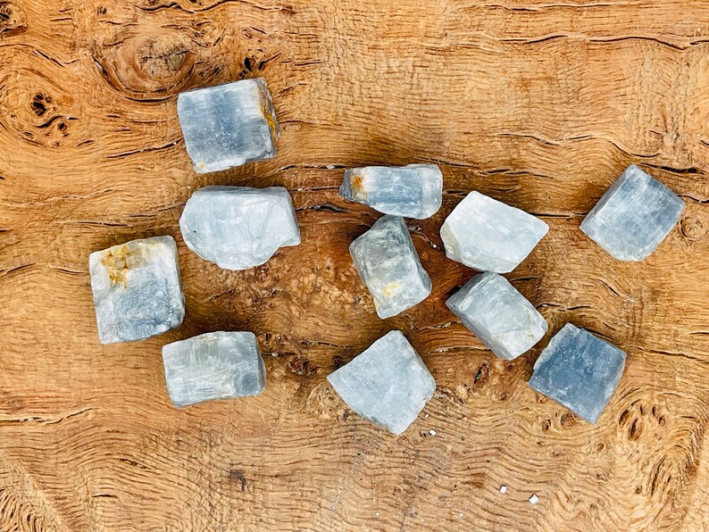 May include: A collection of ten blue calcite crystals, each with a unique shape and texture. The crystals are arranged on a wooden surface.