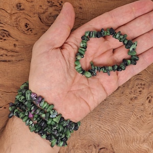 May include: Two beaded bracelets made of green and black gemstone chips, with touches of pink and purple. One bracelet is worn on a wrist, the other in an open hand. The bracelets are shown against a wooden background.