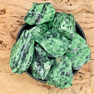 May include: A collection of green and black zultanite rough stones in a black bowl. The stones have a speckled appearance.