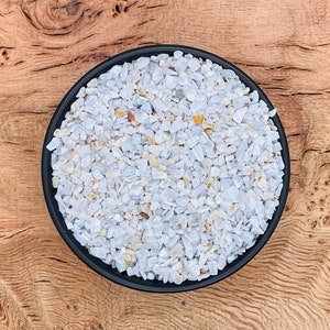 Puede incluir: Un cuenco negro lleno de pequeñas virutas de piedra preciosa pulida de color azul claro y blanco. El cuenco está sobre una superficie de madera.