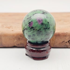 Könnte beinhalten: Eine polierte Edelstein-Kugel, hauptsächlich grün mit dunklen Flecken und rosa Einschlüssen, steht auf einem kleinen, dunkelroten Holzständer. Die Kugel ist glatt und reflektierend.