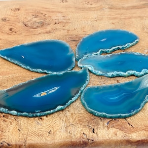May include: Five teal blue agate slices with a natural, irregular shape. The slices are arranged in a starburst pattern on a wooden surface.