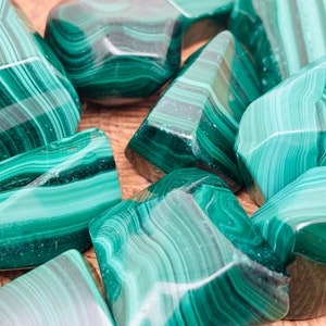 May include: A collection of polished malachite stones. The stones display a rich, deep green color with characteristic swirling patterns. The shapes vary, with some stones appearing more rounded and others more angular. The malachite is displayed on a wooden surface.