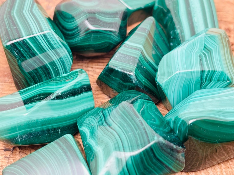 May include: A collection of polished malachite stones. The stones display a rich, deep green color with characteristic swirling patterns. The shapes vary, with some stones appearing more rounded and others more angular. The malachite is displayed on a wooden surface.