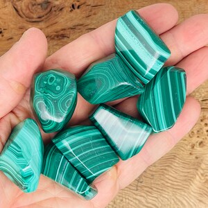May include: A collection of polished malachite stones, each displaying unique patterns of light and dark green bands and swirls. The stones are held in a hand, showcasing their natural beauty and variations in color and texture.