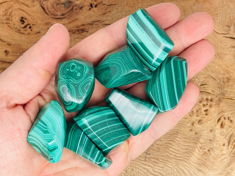 May include: A collection of polished malachite stones, each displaying unique patterns of light and dark green bands and swirls. The stones are held in a hand, showcasing their natural beauty and variations in color and texture.