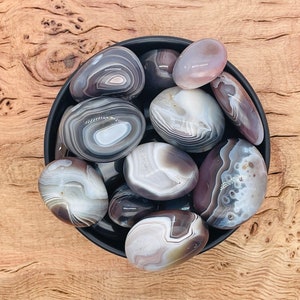 May include: A collection of grey and white banded agate stones in a black bowl. The stones are smooth and polished, with a variety of patterns and textures.