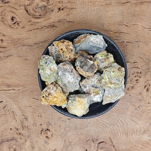 Puede incluir: Un pequeño cuenco lleno de piedras minerales verdes, amarillas y negras. Las piedras son ásperas y tienen un aspecto natural y terroso.
