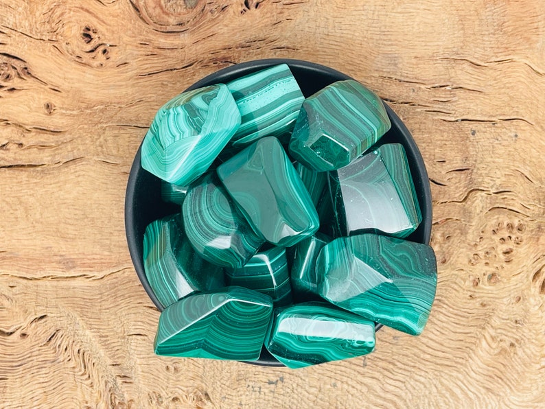 May include: A collection of polished malachite stones in a black bowl. The stones display a vibrant green color with distinctive banded patterns. The bowl sits on a wooden surface, providing a natural backdrop.