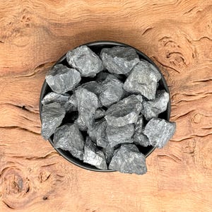 Może przedstawiać: Czarna miska wypełniona kolekcją ciemnoszarych, metalicznych kamieni. Miska stoi na jasnobrązowej drewnianej powierzchni.
