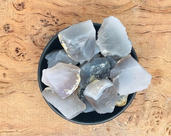 Peut inclure: Une collection de morceaux de cristal d'agate gris et blanc dans un bol noir. Les cristaux ont une surface lisse et polie et sont disposés de manière aléatoire.