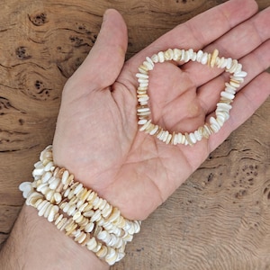 Puede incluir: Un brazalete de concha blanco y beige con múltiples hebras. El brazalete está hecho de pequeñas piezas planas de concha que están enhebradas.
