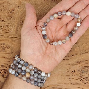 May include: A collection of beaded bracelets in shades of gray, white, and brown. One bracelet is held in an open hand, while several others are stacked on the wrist. The beads are round and appear to be made of stone.
