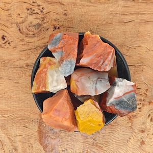 May include: A small black bowl filled with colorful, rough-cut, red, yellow, and white stones. The bowl is sitting on a wooden surface.
